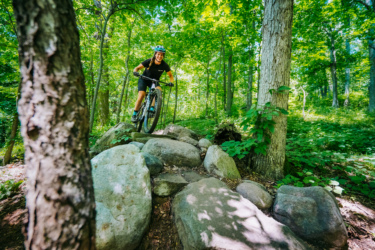 A mountain biker navigating over large rocks on a forest trail, surrounded by lush green trees and foliage. The cyclist is wearing a helmet and a black outfit, showcasing a dynamic ride in a natural setting. DTE Energy Foundation Trails mountain bike trail.