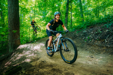 Two mountain bikers navigating a dirt trail through a lush green forest. One biker is riding on a slope with a smile, while the other is performing a jump in the background. The scene is vibrant with sunlight filtering through the trees, highlighting the excitement of the outdoor adventure. DTE Energy Foundation Trails mountain bike trail.