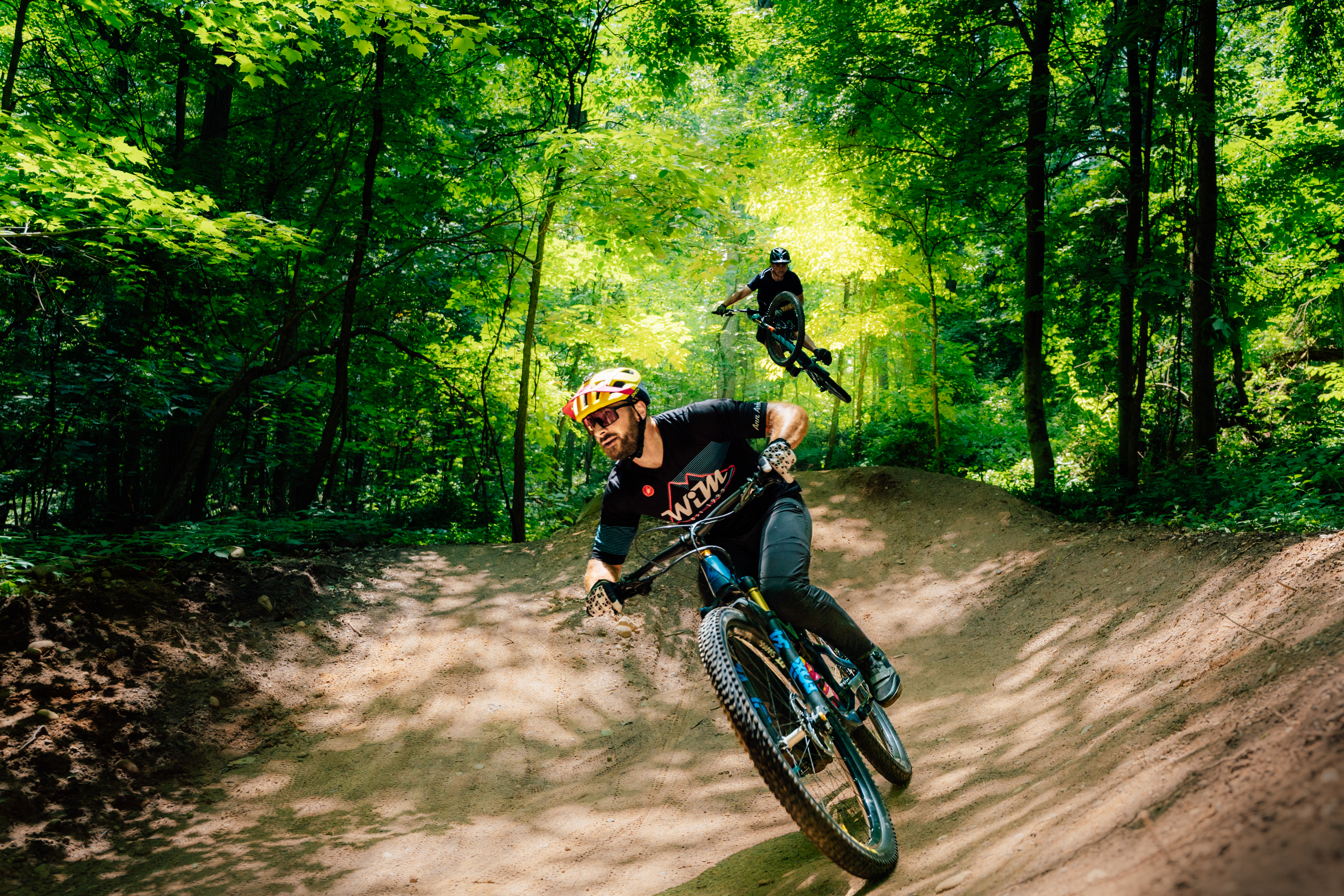 Two mountain bikers navigating a dirt trail in a lush green forest. One biker is speeding around a curve while the other is airborne, performing a jump in the background. Sunlight filters through the trees, creating a vibrant and dynamic outdoor scene. DTE Energy Foundation Trails mountain bike trail.