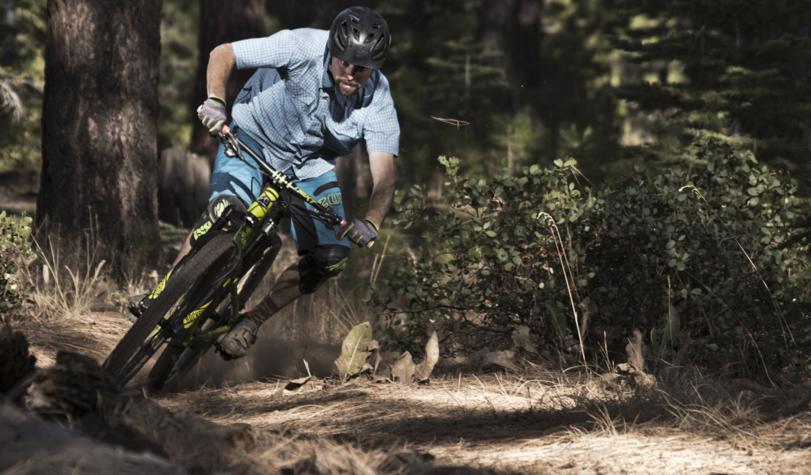 A mountain biker in a blue checkered shirt and patterned shorts leans into a turn on a woodland trail, with trees and greenery surrounding the path. Dust rises from the tires as he navigates the terrain, showcasing an action-packed moment in a natural setting.