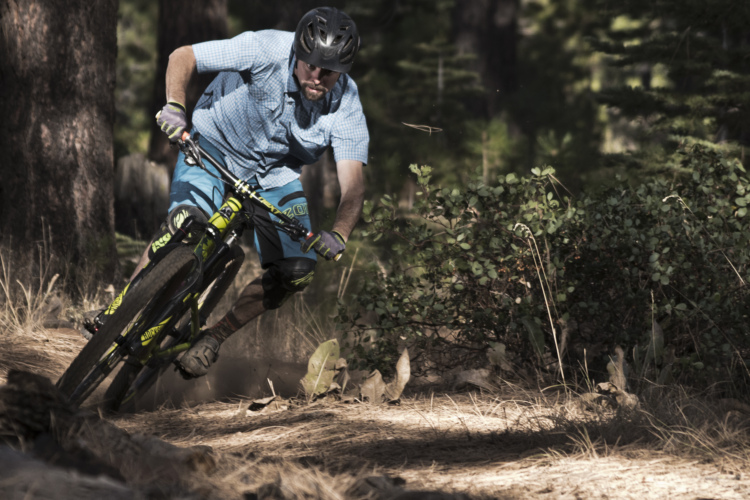 A mountain biker in a blue checkered shirt and patterned shorts leans into a turn on a woodland trail, with trees and greenery surrounding the path. Dust rises from the tires as he navigates the terrain, showcasing an action-packed moment in a natural setting.