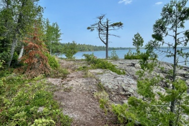 A scenic view of a rocky shoreline surrounded by lush green trees, with a calm lake in the distance under a partly cloudy sky. A narrow path winds through the vegetation, leading towards the water's edge. Keweenaw Point Trail mountain bike trail.