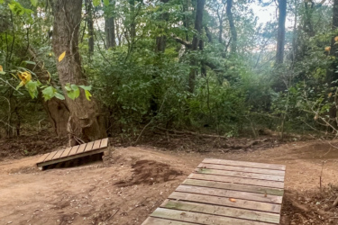 A wooden bridge crossing a narrow path in a dense forest. Surrounding trees and green foliage create a natural, serene environment. The ground is partially covered with dirt and leaves, indicating a well-trodden trail. The Local mountain bike trail.