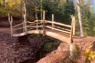 A rustic wooden bridge arching over a small stream, surrounded by trees with autumn foliage. The bridge features a natural design, with logs forming the railing and archway, and is set against a backdrop of vibrant fall colors and leafy underbrush. Keweenaw Point Trail mountain bike trail.