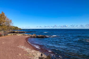 A scenic view of a shoreline with reddish sand and rocky formations, under a clear blue sky with a few clouds. The calm blue water of the lake gently laps at the shore, while trees with autumn foliage line the background. Keweenaw Point Trail mountain bike trail.