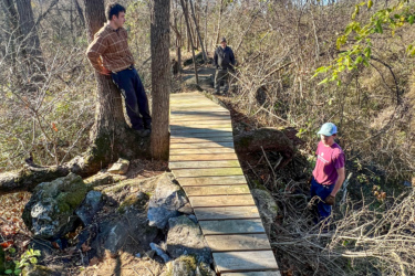 Three individuals are working in a wooded area, overseeing the construction of a wooden bridge that spans over rocks and uneven ground. One person is leaning against a tree, while the others are positioned on the newly constructed pathway surrounded by dense underbrush and trees. The scene captures a moment of outdoor labor and collaboration in a natural setting. The Local mountain bike trail.