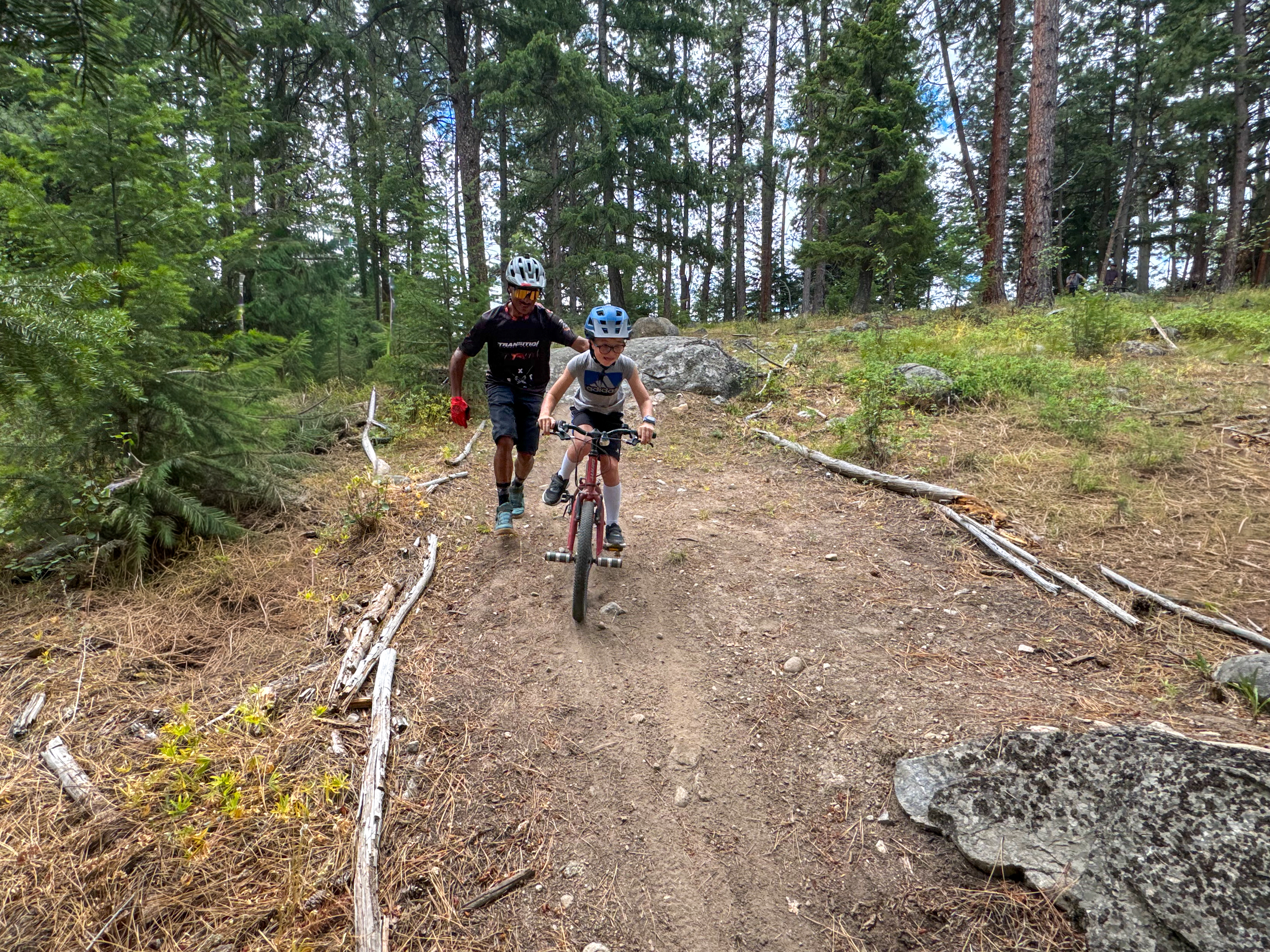 A child riding a bicycle on a dirt trail in a forest, accompanied by an adult who is walking alongside. Both are wearing helmets, and the scene is surrounded by tall trees and natural vegetation. The Property mountain bike trail.