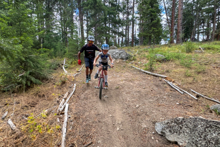 A parent assists a child riding a red bicycle on a dirt trail surrounded by trees and rocks in a forested area. The child wears a helmet and a T-shirt, while the parent encourages them from behind, also wearing a helmet. The scene conveys a sense of outdoor adventure and family bonding.