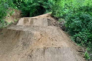 A dirt bike jump area with two ramps surrounded by lush green foliage. The terrain is uneven and earthy, indicating it is used for outdoor biking activities. The Local mountain bike trail.