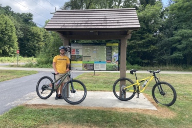 A person wearing a yellow "UT Toledo" t-shirt and a helmet stands next to two mountain bikes at a trailhead sign. The sign displays information about nearby biking trails, and the area is surrounded by trees and grass. Beach Ridge Singletrack mountain bike trail.