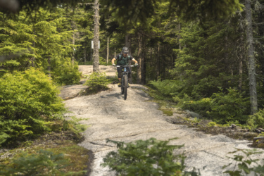 A mountain biker navigating a rocky trail surrounded by lush green trees in a forest setting. The rider is focused and balanced on the bike as they maneuver over the rocky terrain. Slab City mountain bike trail.