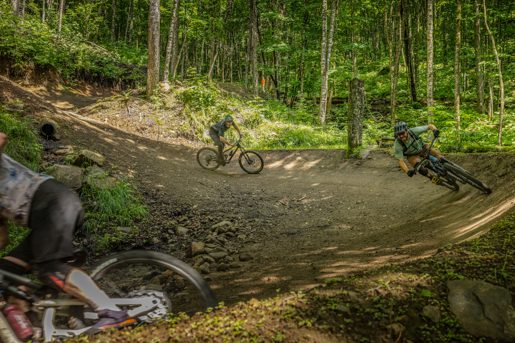 Two mountain bikers navigate a dirt trail in a lush, green forest. One rider is leaning into a turn, showcasing skill and speed, while the other is slightly in the foreground, partially out of focus. The tree-filled background features dappled sunlight filtering through the leaves, creating a vibrant outdoor atmosphere.