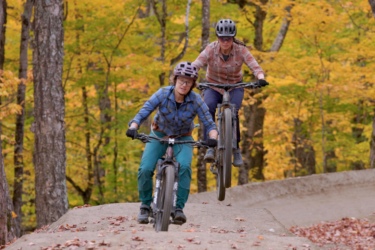 Two mountain bikers riding on a dirt path in a wooded area, surrounded by vibrant autumn foliage. One rider is performing a jump while the other is focused on navigating the terrain. Both are wearing helmets and casual cycling attire. Sugarloaf Bike Park mountain bike trail.