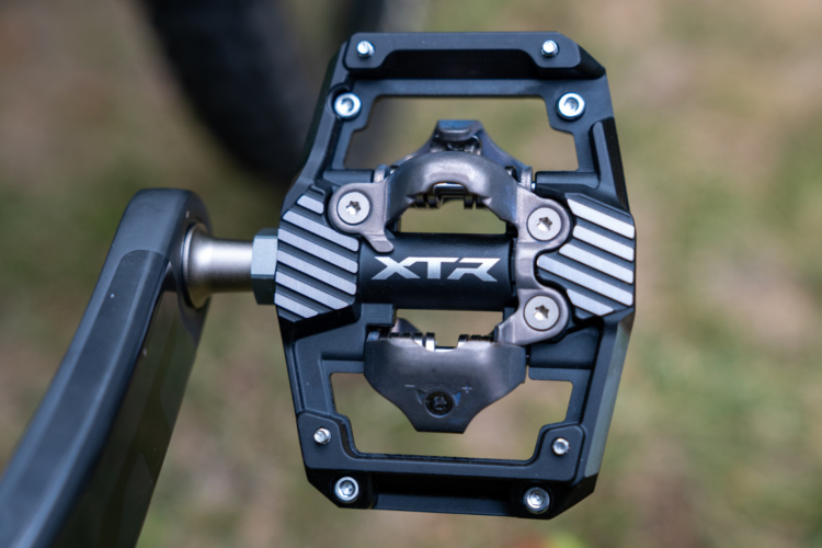 Close-up of a Shimano XTR bicycle pedal, featuring a sleek metal and black design with a prominent XTR logo, mounted on a crank arm. The background shows a blurred outdoor setting.