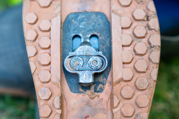Close-up view of a bicycle cleat attached to the sole of a cycling shoe, featuring a metallic connector plate and two screws, set against a textured rubber sole with a hexagonal pattern.