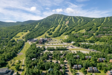 Aerial view of a mountainous landscape featuring a ski resort with green slopes, surrounded by dense forests. In the foreground, various buildings and parking areas are visible, with multiple residential structures located at the base of the mountain. The sky is partly cloudy, and ski trails can be seen marked on the mountainside. Sugarloaf Bike Park mountain bike trail.