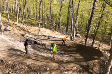 Three individuals are working on a dirt biking trail in a forested area. They are using shovels and rakes to shape the terrain, creating dips and features in the dirt. Surrounding them are trees with fresh green leaves, indicating a spring setting. Tools and materials are scattered nearby, and the atmosphere appears focused and industrious. Sugarloaf Bike Park mountain bike trail.