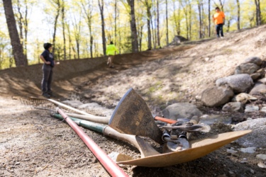 A close-up view of gardening tools including a shovel, rake, and pruners arranged on the ground, with a blurred background showing two people working on a trail in a wooded area filled with green trees. Sugarloaf Bike Park mountain bike trail.