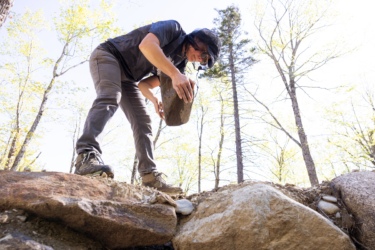 A person in outdoor gear is lifting a large rock while balancing on a rocky surface in a wooded area with trees in the background. The scene is bright and sunny, suggesting an active outdoor activity, possibly related to landscaping or construction. Sugarloaf Bike Park mountain bike trail.
