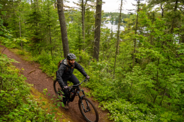 A mountain biker riding along a narrow, dirt trail surrounded by lush green trees and vegetation. The cyclist wears a helmet and is focused on the path ahead, with a serene lake visible in the background. Keweenaw Point Trail mountain bike trail.