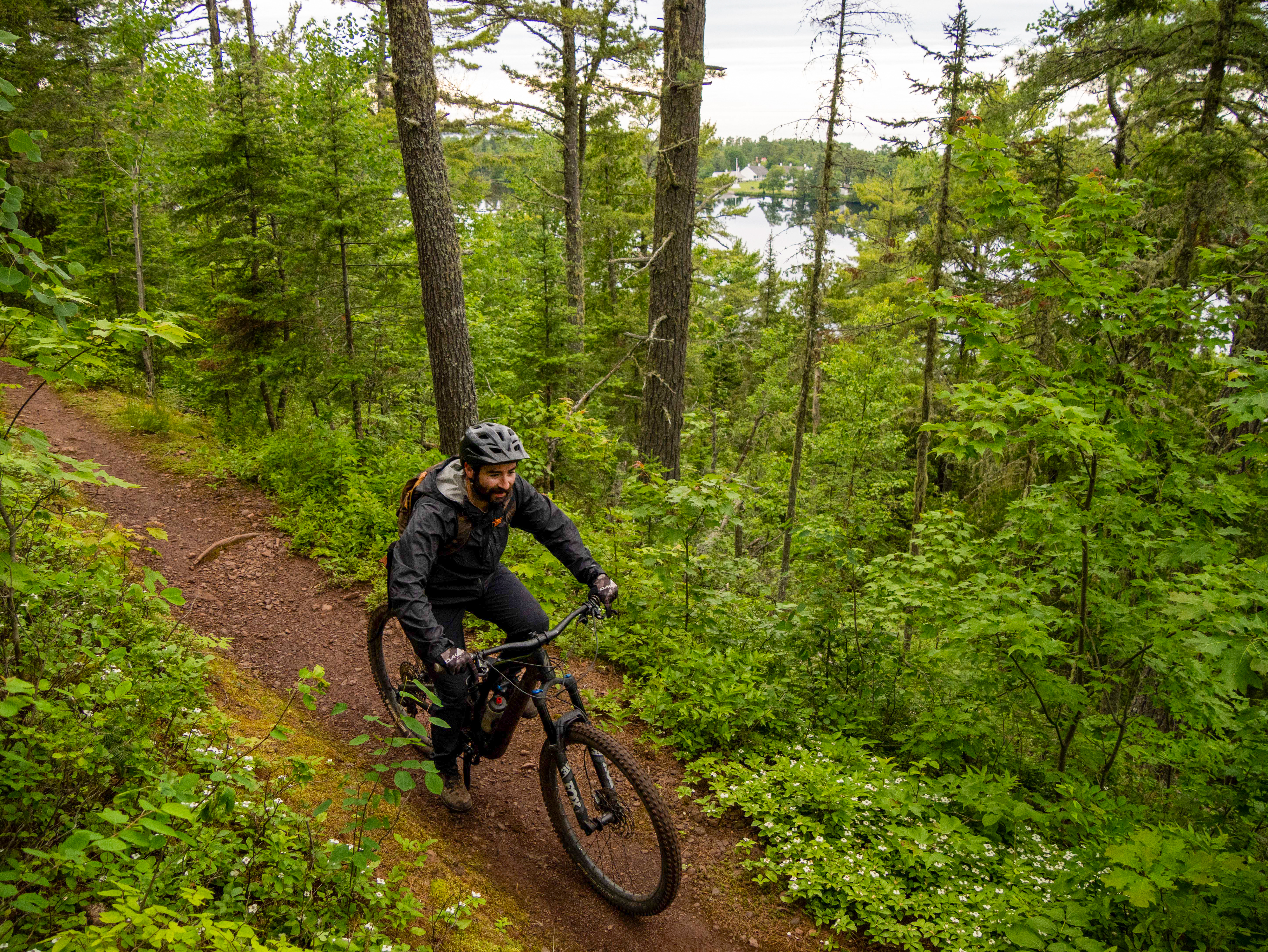 A mountain biker riding along a narrow, dirt trail surrounded by lush green trees and vegetation. The cyclist wears a helmet and is focused on the path ahead, with a serene lake visible in the background. Keweenaw Point Trail mountain bike trail.
