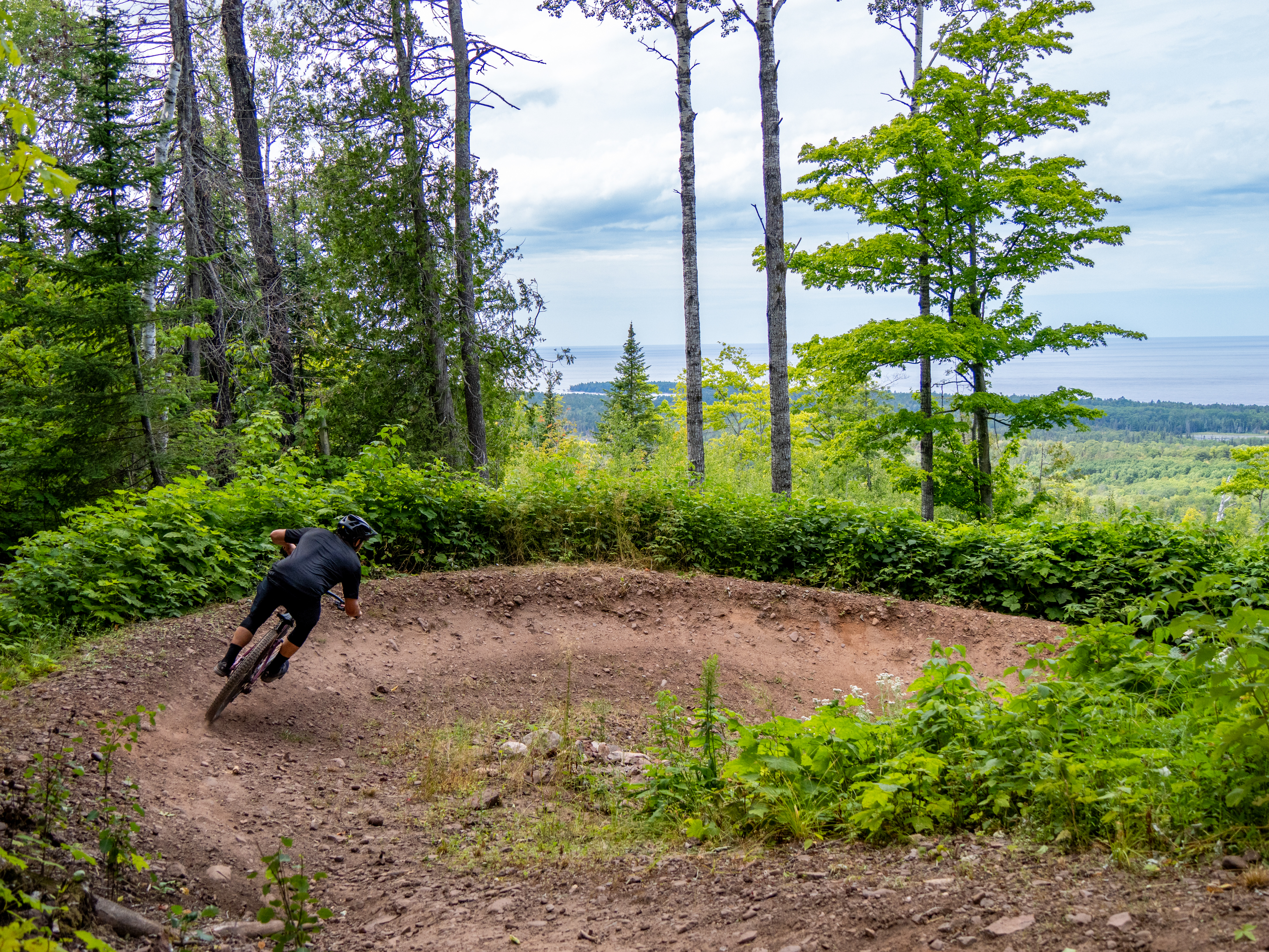A mountain biker navigating a dirt trail surrounded by lush greenery and tall trees, with a scenic view of hills and a body of water in the background. The biker leans into a curve on the trail, showcasing an action shot of the ride. East Bluff Bike Park mountain bike trail.