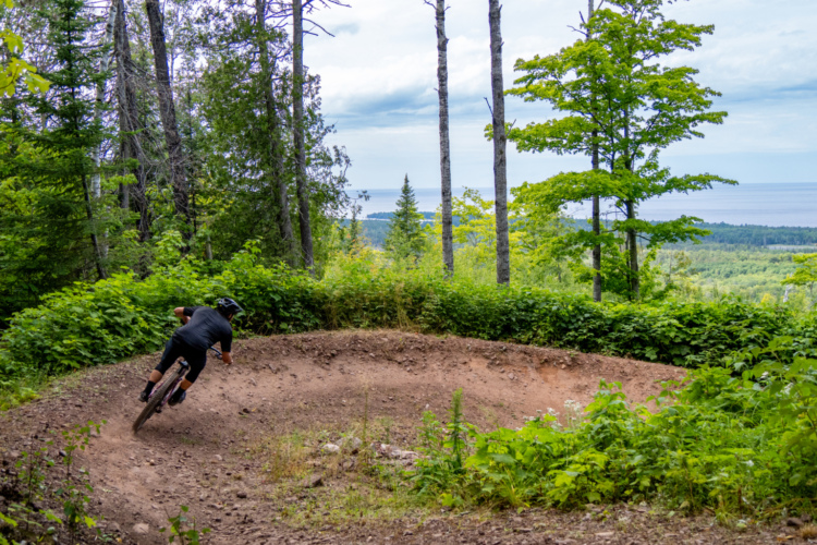A mountain biker navigating a curved trail through a lush green forest, with tall trees and a scenic view of a lake in the distance. The rider is leaning into the turn, showcasing the dynamic action of mountain biking.