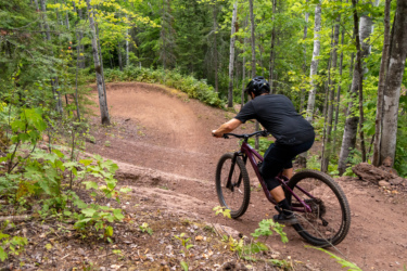 A mountain biker rides along a dirt path through a lush green forest, navigating a curve in the trail surrounded by trees and foliage. East Bluff Bike Park mountain bike trail.