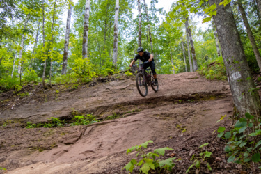 A mountain biker navigating a steep, muddy incline in a forested area, surrounded by tall trees and green foliage. East Bluff Bike Park mountain bike trail.