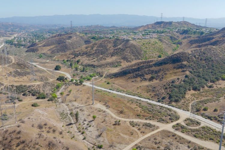 Aerial view of rolling hills and valleys in a dry landscape, intersected by winding dirt paths and a paved road. Vegetation is sparse, with patches of greenery visible among the brown terrain. Power lines stretch across the hills, and a suburban neighborhood is visible in the distance, framed by mountains under a clear blue sky.