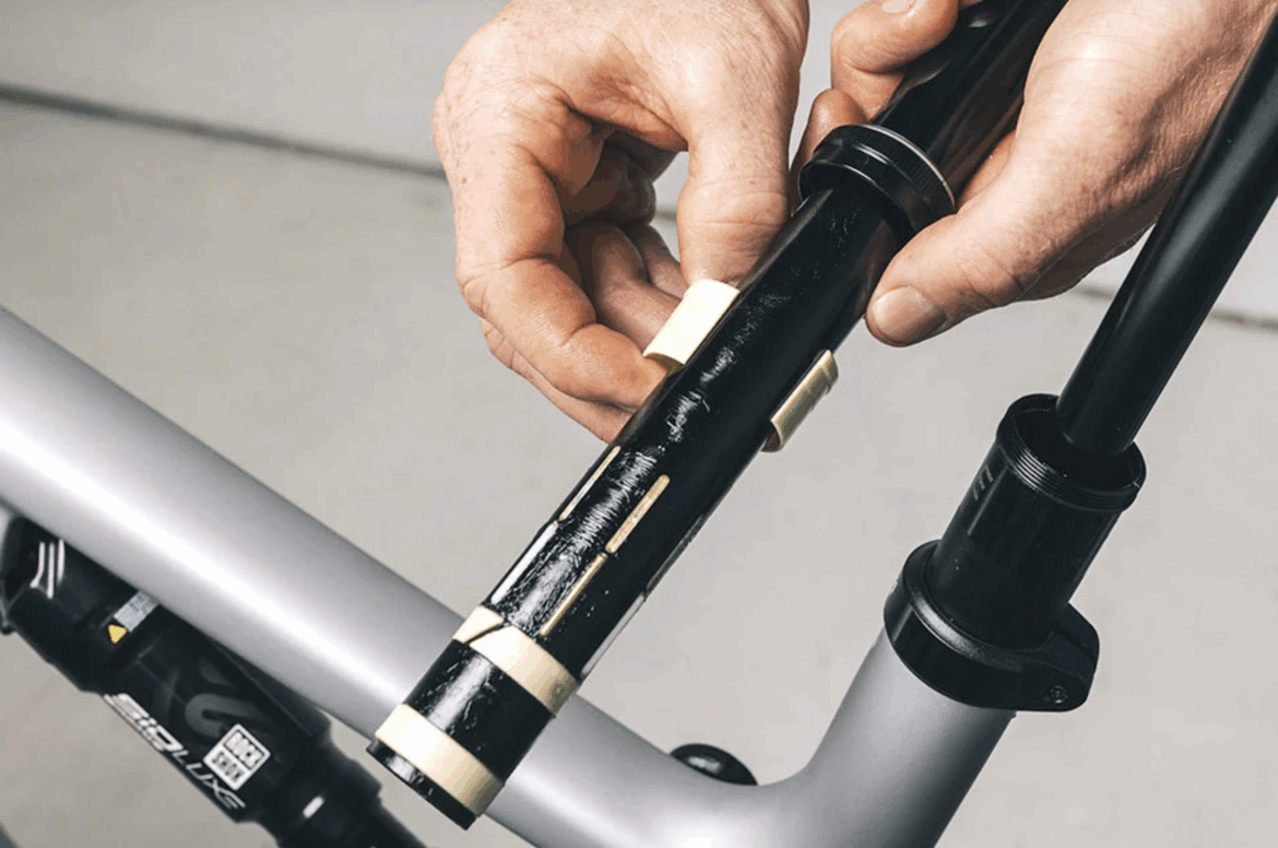 A person’s hands are shown adjusting a black component on a bicycle frame. The component has markings and small attachments, and is being fitted into a part of the bike's structure. The background is a neutral color, emphasizing the focus on the bicycle mechanics.