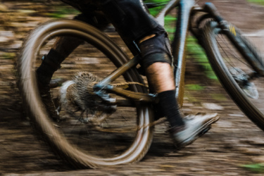 A close-up of a mountain bike's rear wheel in motion, with mud splattering as the bike navigates a dirt trail. The cyclist's leg and lower body are partially visible, wearing black shorts and socks. The surrounding environment is blurred, emphasizing the speed and dynamic action of the ride. Franknbeans mountain bike trail.