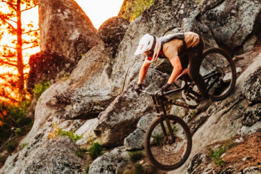 A mountain biker navigating rocky terrain while descending a steep slope during sunset, surrounded by trees and boulders. The rider is wearing a helmet and has a focused expression as they maneuver over the rugged landscape. Hobo Juice mountain bike trail.