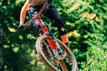 A young cyclist wearing a helmet and protective gear performs a jump on a mountain bike, flying through the air with green trees in the background. Dust and grass are kicked up from the landing, capturing a dynamic moment in a mountain biking scene. Monumental mountain bike trail.