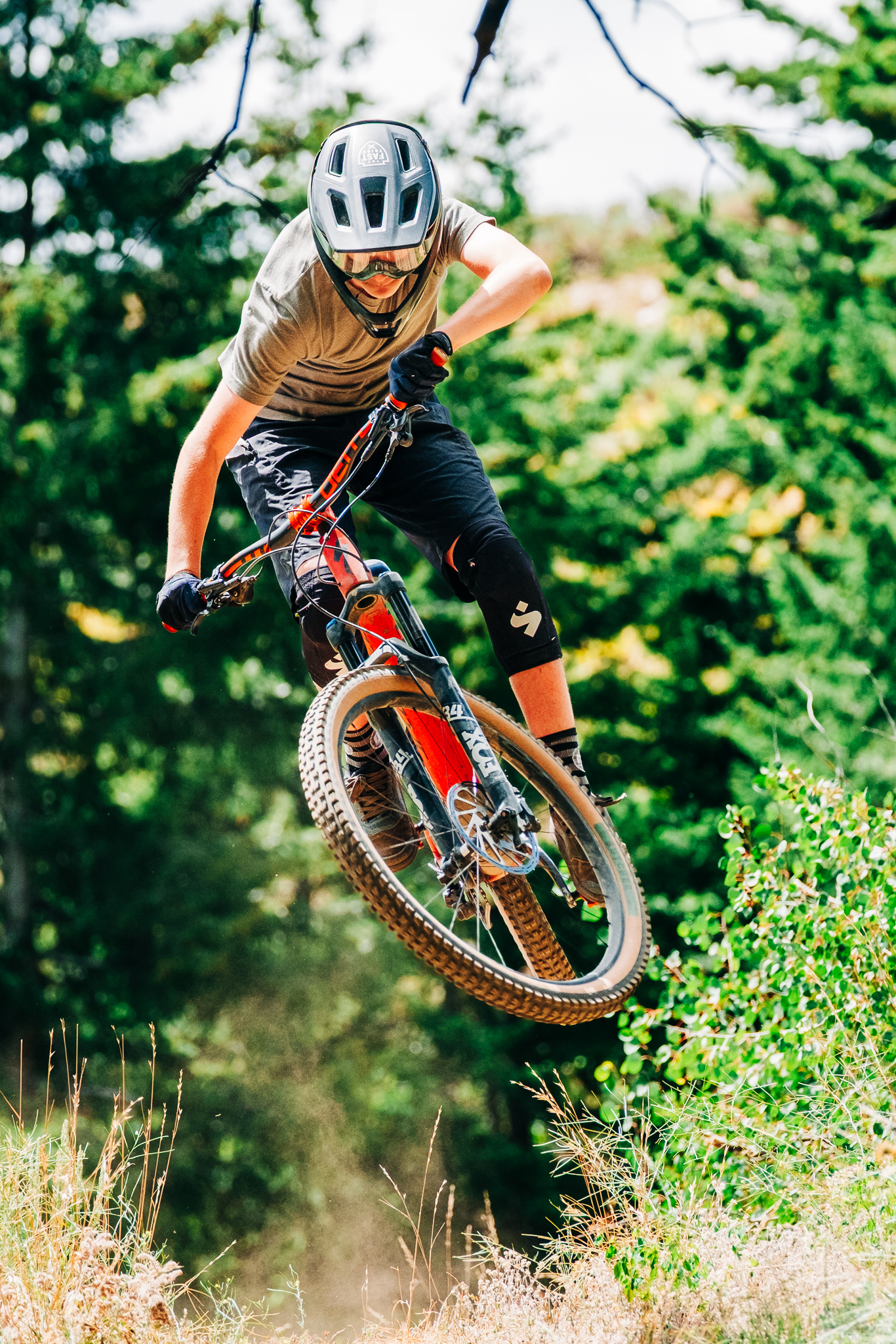 A young cyclist wearing a helmet and protective gear performs a jump on a mountain bike, flying through the air with green trees in the background. Dust and grass are kicked up from the landing, capturing a dynamic moment in a mountain biking scene. Monumental mountain bike trail.