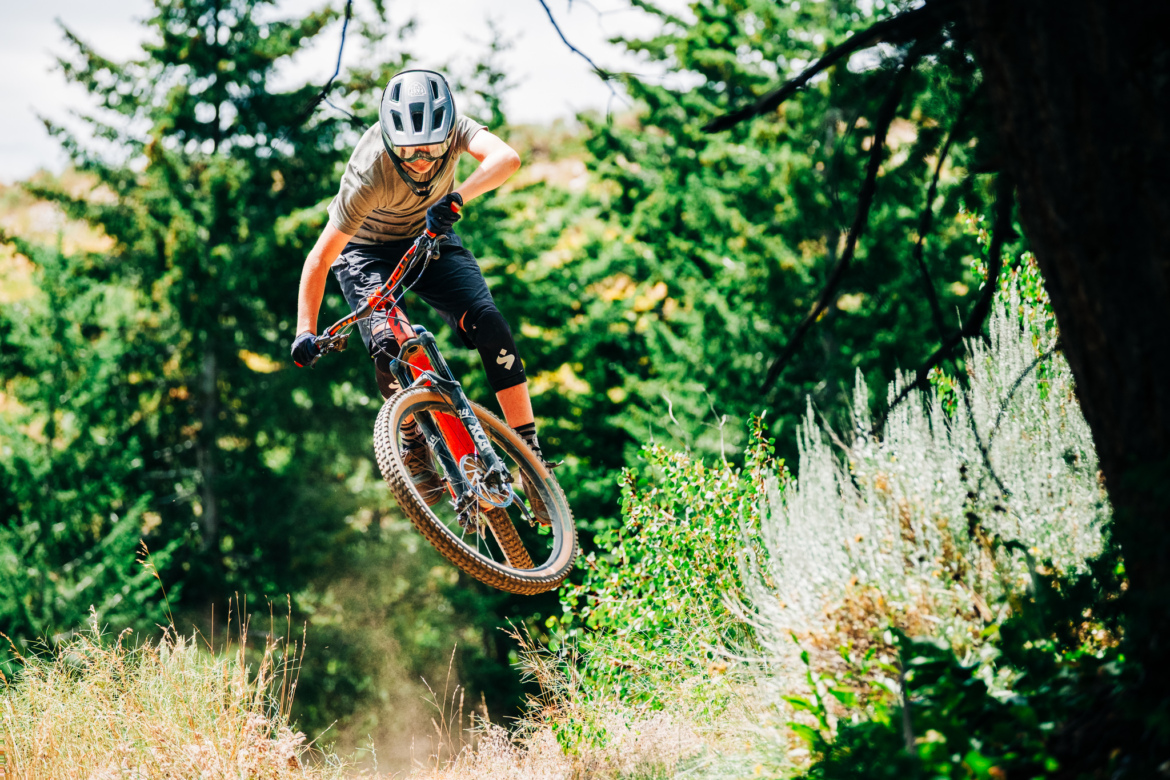 A young mountain biker performing a jump on a trail surrounded by trees and greenery, wearing a helmet and protective gear. Dust is kicked up from the ground as the bike soars through the air.