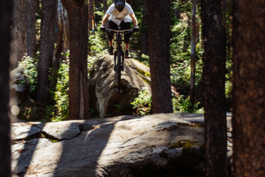 A cyclist in a black helmet and white shirt is airborne over a large rock in a sunlit forest. Tall trees surround the area, providing a natural trail backdrop with patches of sunlight filtering through the leaves. Pura Vida mountain bike trail.