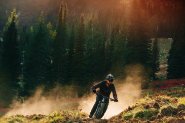 A mountain biker navigating a dirt trail through a forested landscape, with dust rising around him as the sun sets behind the trees, creating a warm, golden light. High Point mountain bike trail.