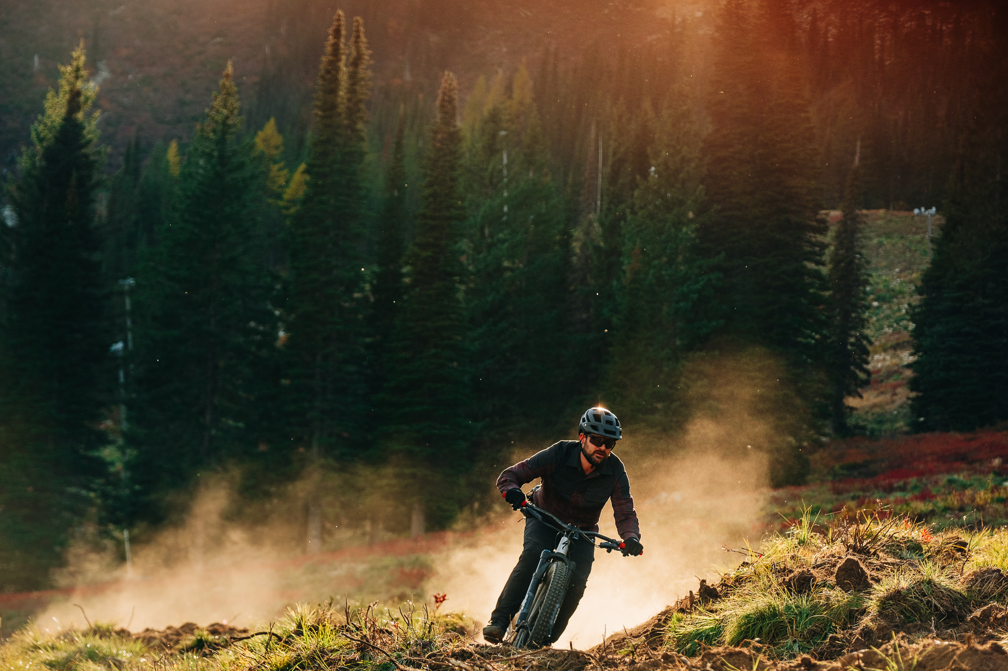 A mountain biker navigating a dirt trail through a forested landscape, with dust rising around him as the sun sets behind the trees, creating a warm, golden light. High Point mountain bike trail.