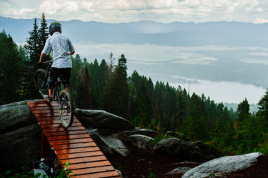 A mountain biker navigates a wooden bridge on a rocky trail, surrounded by lush green trees and a scenic valley below, under a cloudy sky. Smoke Jumper mountain bike trail.