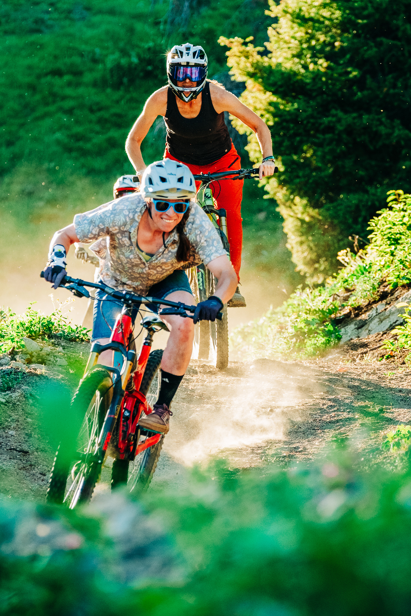 Two mountain bikers riding on a dirt trail, surrounded by greenery. The first rider, wearing a helmet and sunglasses, leans forward on a red bike as dust kicks up behind her. The second rider follows closely, dressed in a black tank top and red pants, navigating the trail uphill. The scene captures a dynamic moment in outdoor cycling amidst a sunny backdrop. Student Rider mountain bike trail.