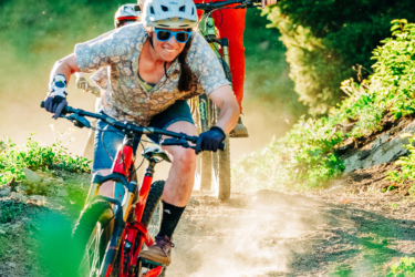 Two mountain bikers navigate a dusty trail surrounded by greenery. The first rider, wearing sunglasses and a helmet, leans forward on a red bike, showcasing a focused expression. The second rider, dressed in a black tank top and red pants, is slightly behind, also on a bike, with sunlight filtering through the trees in the background. Dust is kicked up from the trail, adding to the dynamic feel of the scene. Smoke Jumper mountain bike trail.
