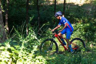 A young cyclist in a blue and red cycling jersey navigates a mountain bike through a lush, green forest trail, surrounded by trees and dense vegetation. The sunlight filters through the foliage, creating a scenic outdoor atmosphere. Federer’s Folly mountain bike trail.