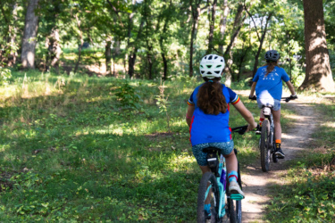 Two young girls ride their bicycles along a winding dirt path through a wooded area, surrounded by greenery and sunlight filtering through the trees. One girl wears a blue shirt and patterned shorts, while the other is in a bright blue shirt with colorful sleeves and a helmet. Federer’s Folly mountain bike trail.