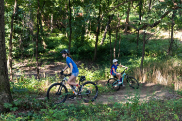Two children riding mountain bikes along a dirt trail in a wooded area, surrounded by greenery and trees. The older child is wearing a helmet and a blue shirt, while the younger child is in a helmet with a colorful outfit. Sunlight filters through the trees, creating a vibrant outdoor scene. Federer’s Folly mountain bike trail.