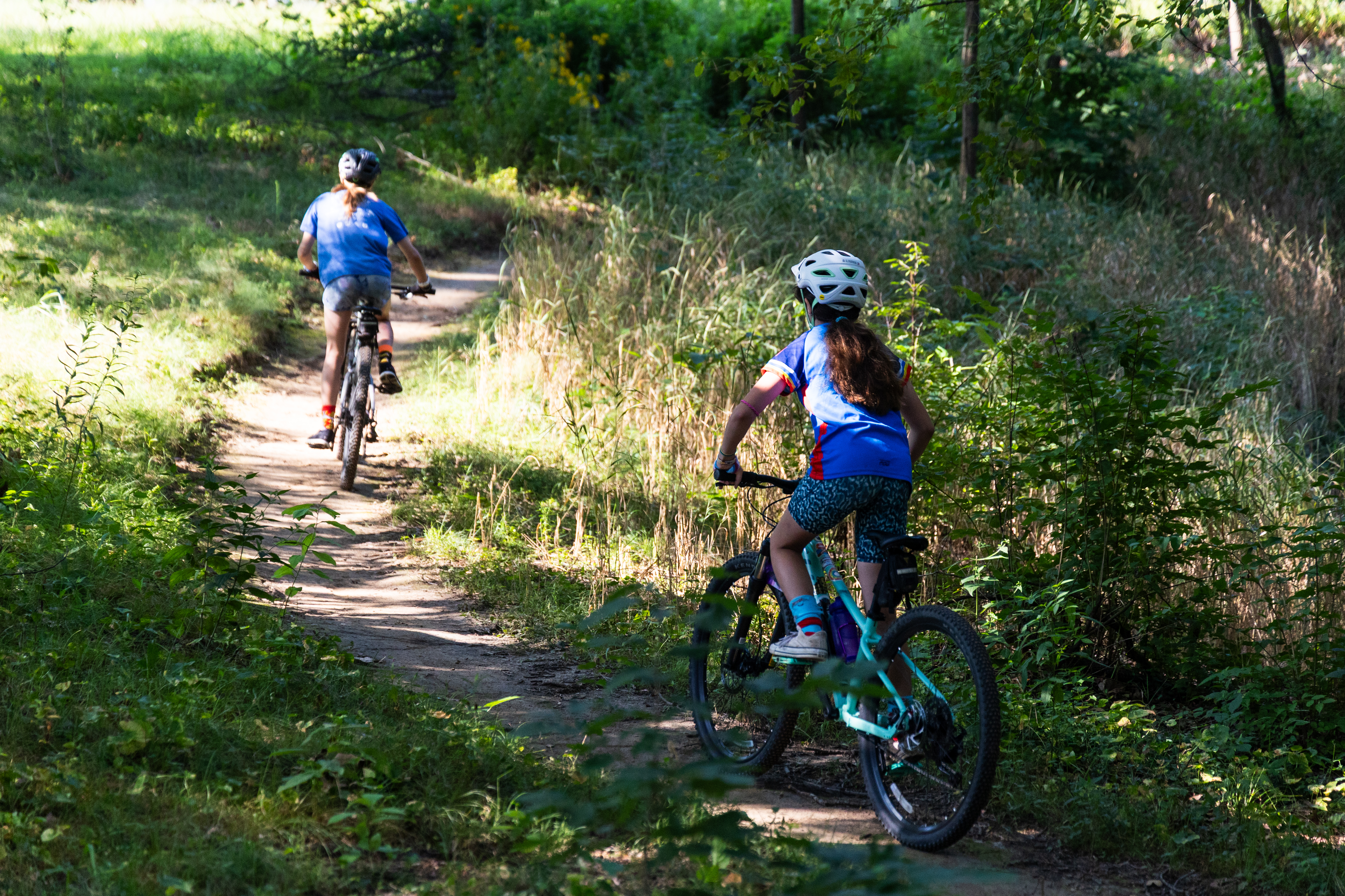 Two children riding bicycles on a winding dirt path through a lush, green forested area. One child is wearing a blue shirt and a helmet while the other wears a blue and red shirt. Sunlight filters through the trees, creating a bright, inviting atmosphere. Federer’s Folly mountain bike trail.