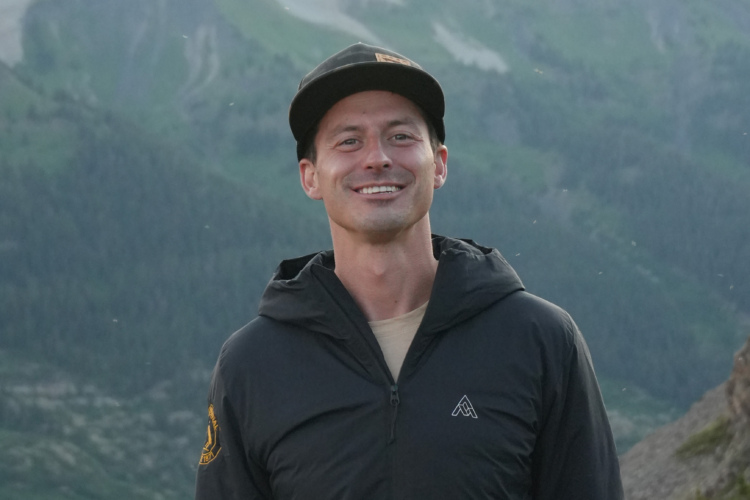 A smiling person wearing a black jacket and cap stands in front of a mountainous landscape with green hills and trees in the background.