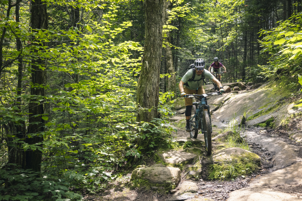 Mountain bikers navigate a rocky trail surrounded by lush green foliage in a wooded area. One cyclist is seen in the foreground, leaning forward as they ride over smooth stones, while another cyclist is slightly behind, making their way through the trail. Sunlight filters through the trees, highlighting the vibrant greenery of the forest.