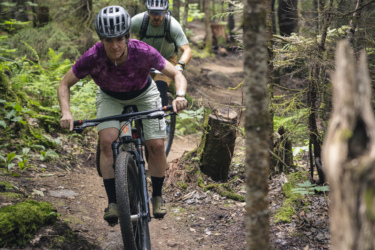 Two mountain bikers navigate a narrow dirt trail through a dense forest. The person in front is focused on the path ahead, wearing a purple shirt and shorts, while the second biker follows closely behind, dressed in a green shirt and equipped with a backpack. Surrounding them are lush greenery and trees, creating a vibrant and natural atmosphere. Mont-Sainte-Anne mountain bike trail.