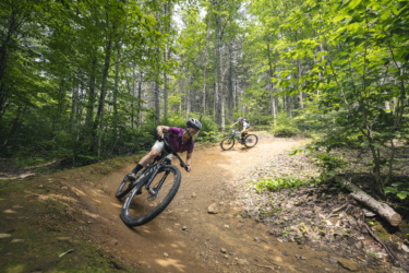 Two mountain bikers navigate a dirt trail through a lush green forest. One rider leans into a curve, while the other follows closely behind. Sunlight filters through the trees, highlighting the vibrant foliage surrounding the trail. Mont-Sainte-Anne mountain bike trail.